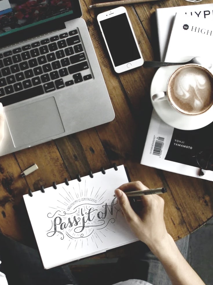 Top down of someone working at a cafe table with a notepad,
        laptop, smartphone, drawing supplies, and a cappuccino with beautiful
        latte art resting on top of a stack on magazines.