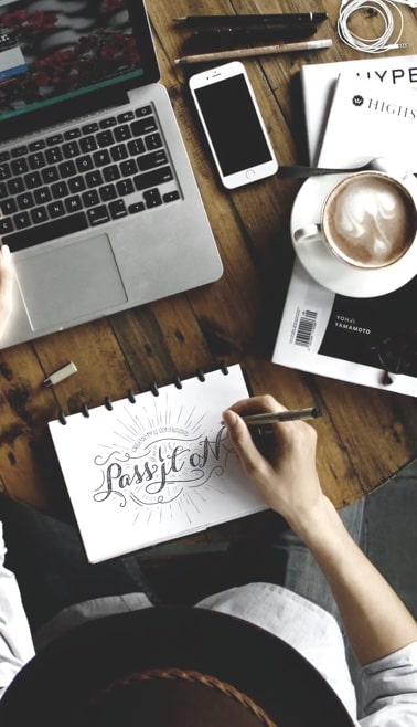 Top down of someone working at a cafe table with a notepad,
        laptop, smartphone, drawing supplies, and a cappuccino with beautiful
        latte art resting on top of a stack on magazines.